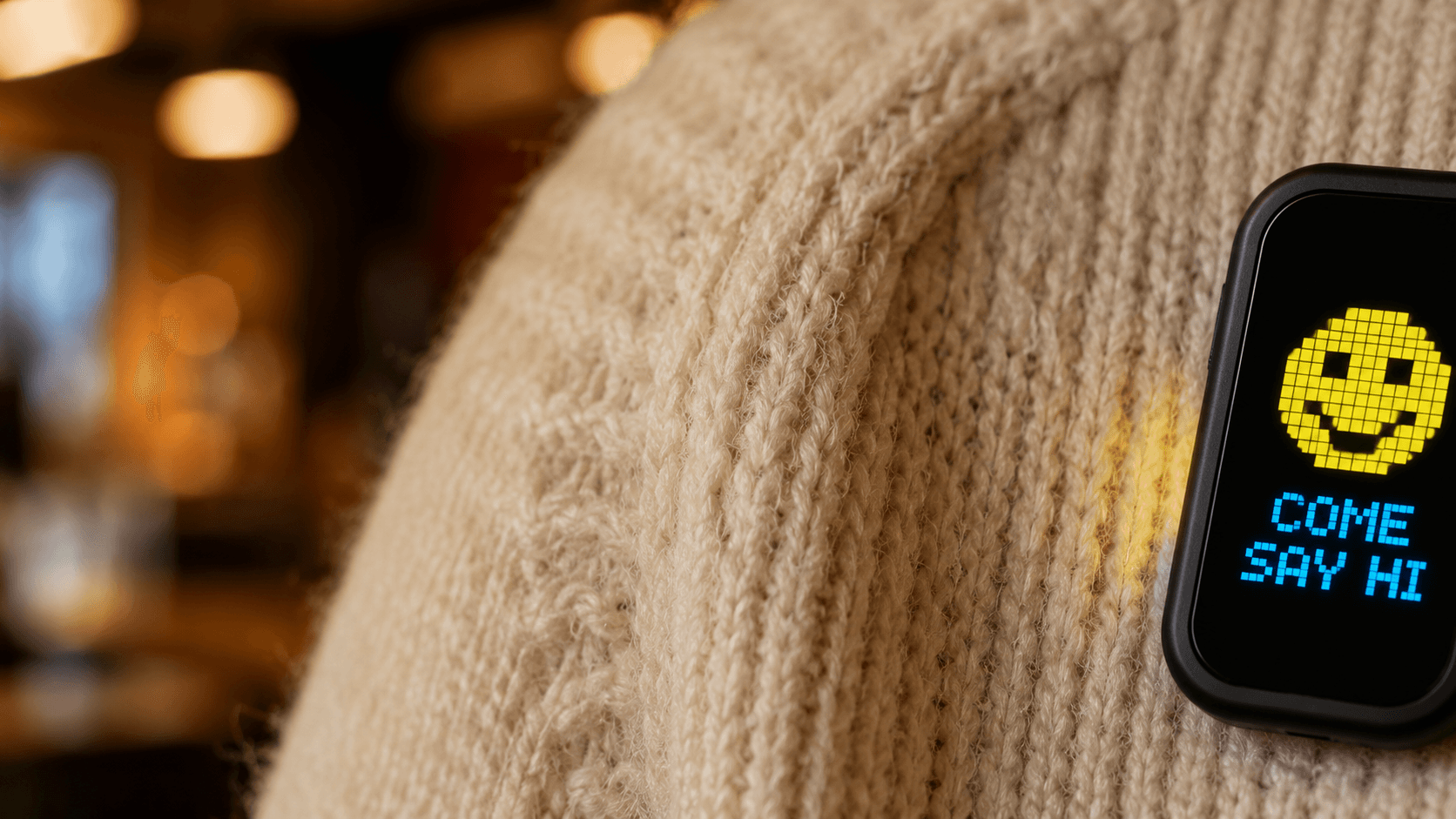 Close-up of a MoodPin on a knit sweater showing a pixel smiley and the words COME SAY HI
